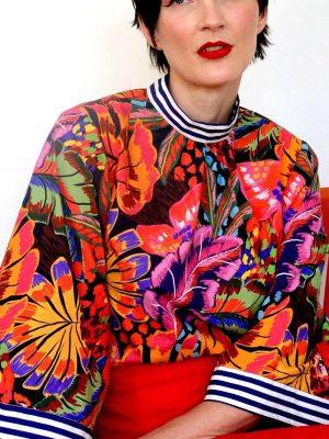 Bold tropical floral blouse featuring vibrant orange, purple, and green hues, paired with a red skirt.