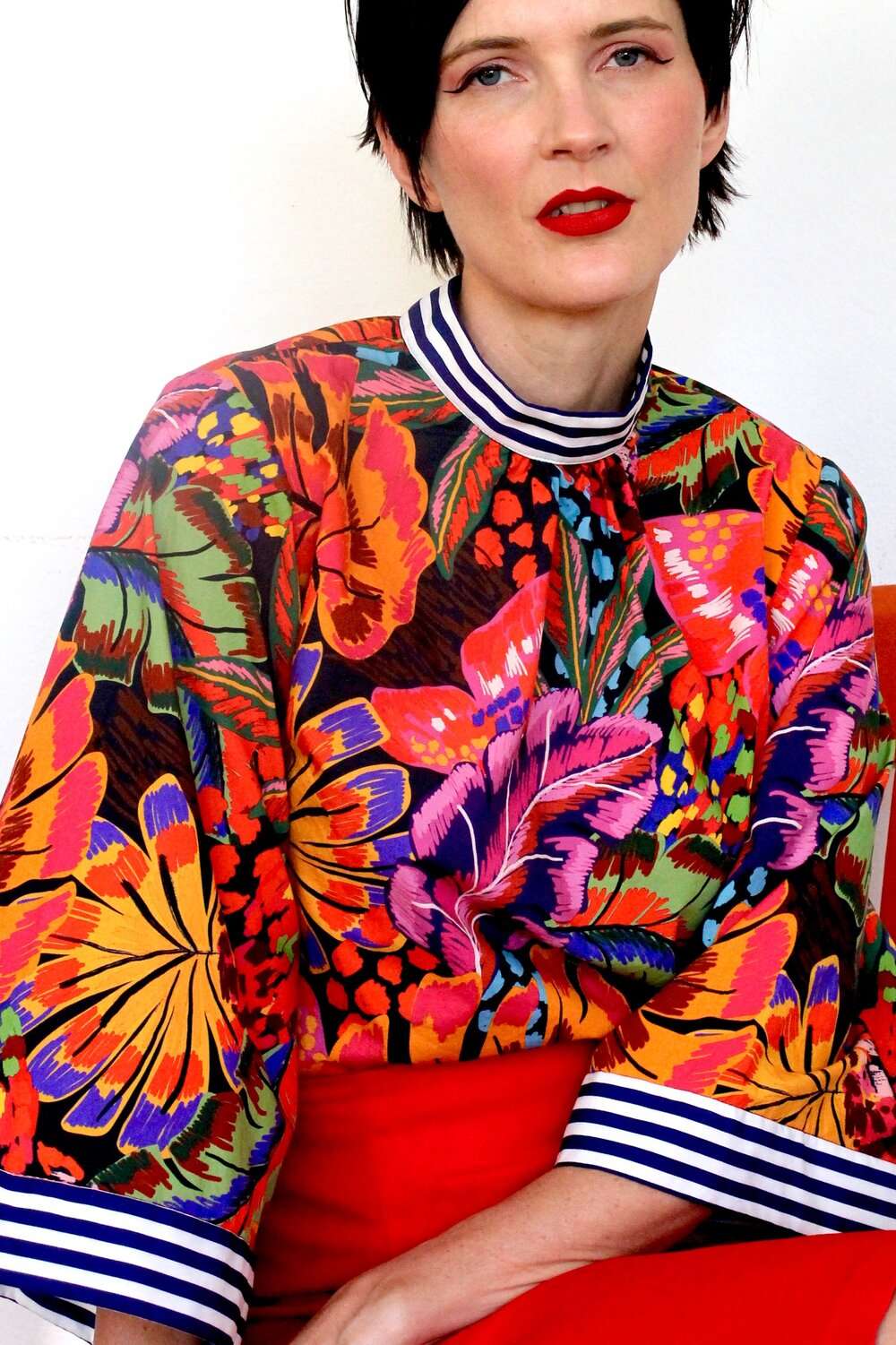 Bold tropical floral blouse featuring vibrant orange, purple, and green hues, paired with a red skirt.