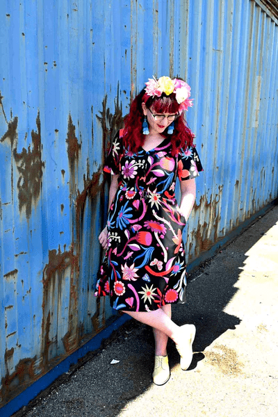 Woman dressed in a black dress with large, bold floral designs in reds, blues, and oranges, styled with a flower crown and leaning casually against a weathered blue wall. Designed by Australian designer brand Streborclothing.