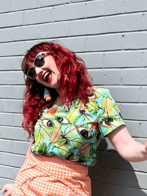 A short-sleeved top with a bold leaf and seed pattern in vibrant green, orange, and black on a light blue background, displayed against a pink backdrop to highlight its colorful appeal. Designed by Australian designer brand Streborclothing.