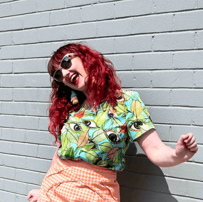 A short-sleeved top with a bold leaf and seed pattern in vibrant green, orange, and black on a light blue background, displayed against a pink backdrop to highlight its colorful appeal. Designed by Australian designer brand Streborclothing.