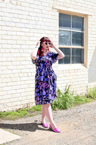 Woman wearing a dark dress with a whimsical cactus and desert-themed print in pinks, purples, and blues, standing against a graphic backdrop with matching patterns. Designed by Australian designer brand Streborclothing.
