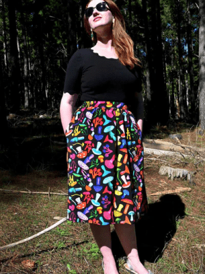 The skirt features a vibrant mushroom pattern with various brightly colored mushrooms in red, yellow, blue, pink, and green on a black background. This playful design adds a whimsical and organic feel to the outfit. Designed by Australian designer brand Streborclothing.