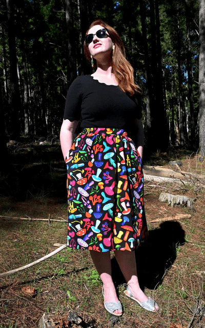 The skirt features a vibrant mushroom pattern with various brightly colored mushrooms in red, yellow, blue, pink, and green on a black background. This playful design adds a whimsical and organic feel to the outfit. Designed by Australian designer brand Streborclothing.