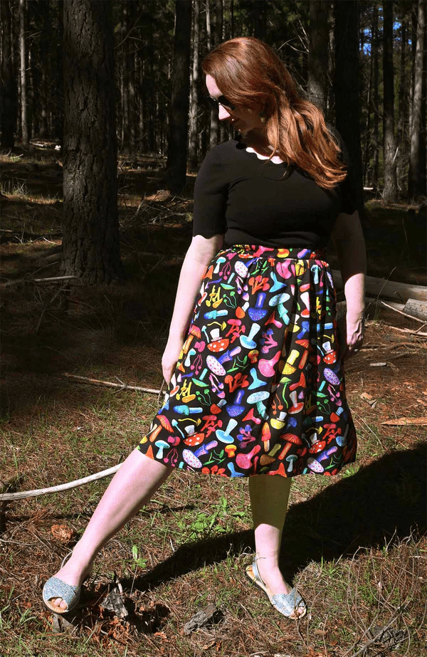 The skirt features a vibrant mushroom pattern with various brightly colored mushrooms in red, yellow, blue, pink, and green on a black background. This playful design adds a whimsical and organic feel to the outfit. Designed by Australian designer brand Streborclothing.