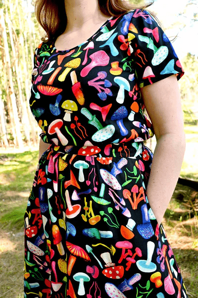 The dress features a vibrant mushroom pattern with various brightly colored mushrooms in red, yellow, blue, pink, and green on a black background. This playful design adds a whimsical and organic feel to the outfit. Designed by Australian designer brand Streborclothing.
