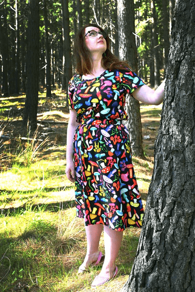 The dress features a vibrant mushroom pattern with various brightly colored mushrooms in red, yellow, blue, pink, and green on a black background. This playful design adds a whimsical and organic feel to the outfit. Designed by Australian designer brand Streborclothing.